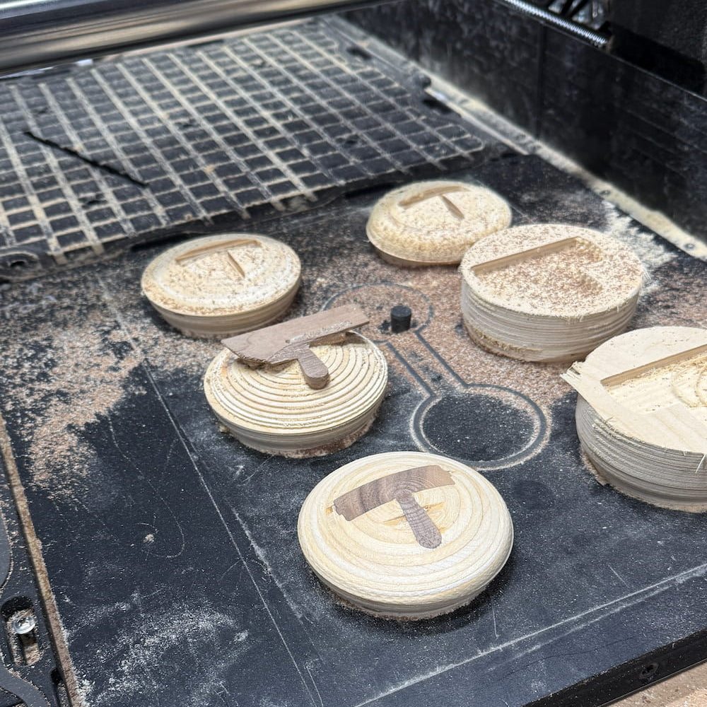 Several unfinished light wood oval plaques or trays with circular grooved details are arranged on a dark work surface with sawdust, near a metal grate and cutting equipment.